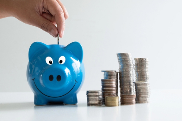 hand putting coin inside a blue piggy bank beside piles of coins